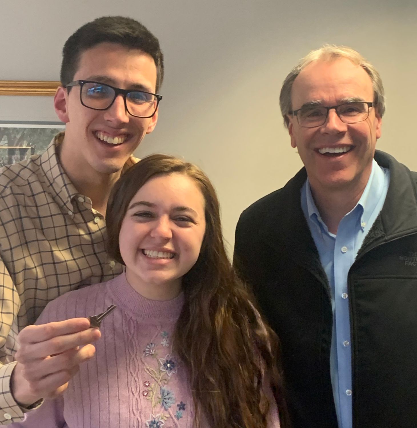 Two happy clients - a man and woman - stand next to David Campbell, Realtor - all smiling and facing towards the camera. The man is holding a house key.
