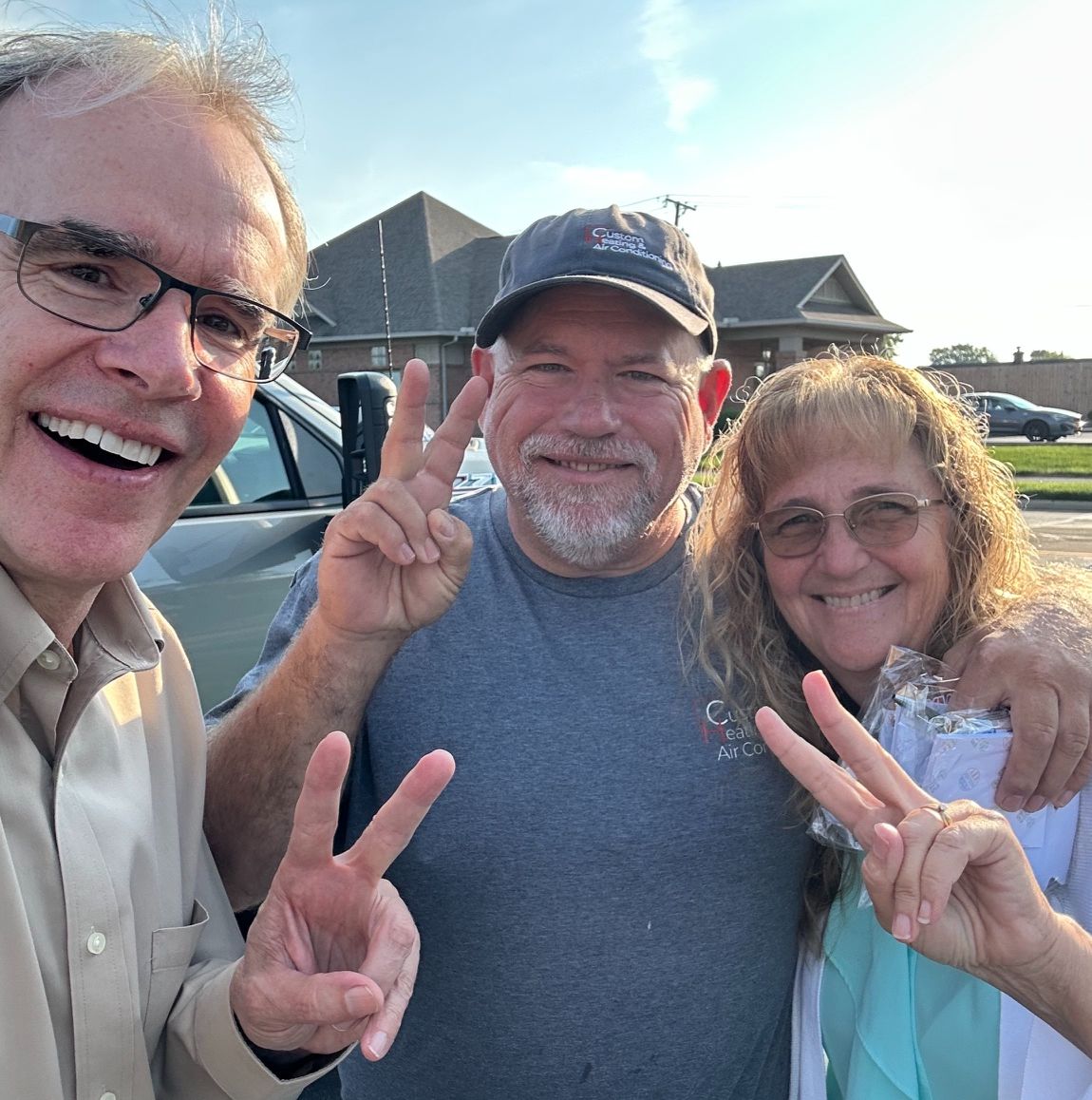 David Campbell, realtor stands next to both Dwayne and Laura. All three are facing the camera while holding up two fingers.