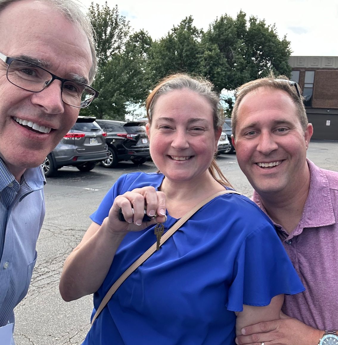 Joe and Amanda stand next to David Campbell, realtor in a parking lot. They're all smiling and Amanda is holding house keys.