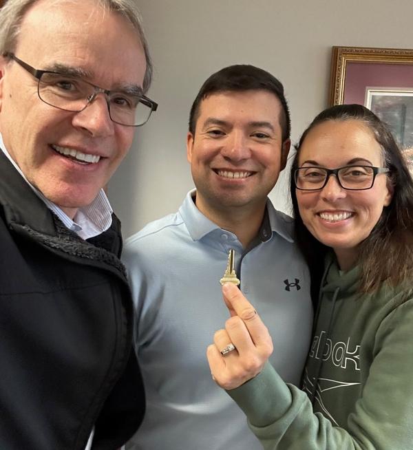 David Campbell, realtor, stands next to both Carlos and Angela as they all face towards the camera, smiling. Angela is holding up a house key.