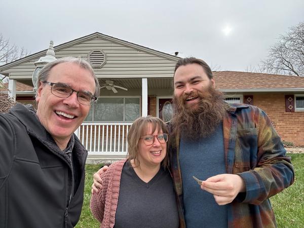 David Campbell stands smiling next to a man and woman. All are smiling and facing towards the camera while the man holds up a house key.