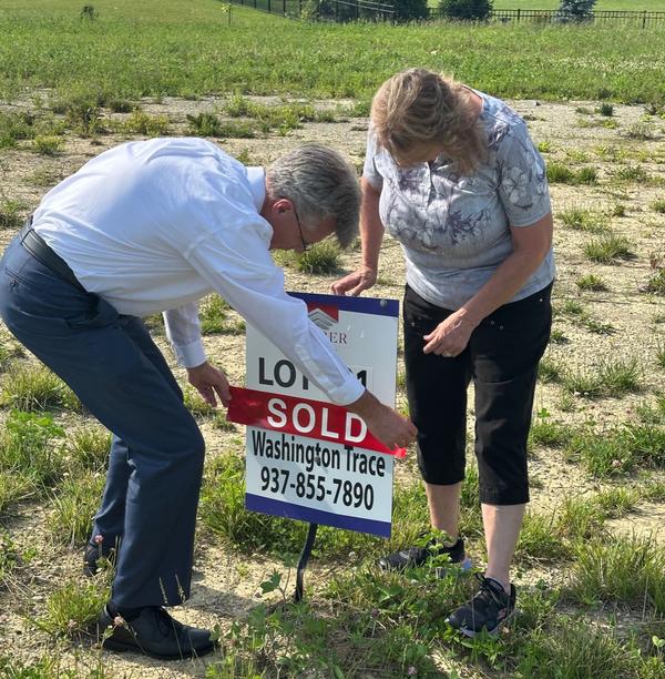 David Campbell, Realtor leans over to put a SOLD banner over a real estate sign next a woman, who's watching him put it in place.
