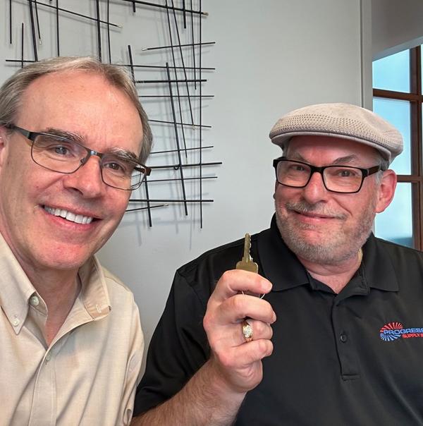 David Campbell, Realtor sits next to a man. They're both looking towards the camera, smiling - the man is holding a house key after a successful home sale.