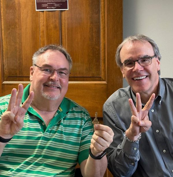 David Campbell, realtor, sits next to a man, Mark, while they both face towards the camera, smiling and holding up three fingers each. Mark is holding a house key in his left hand.
