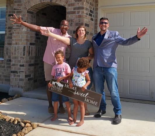 Smiling family of four stands barefoot with Anthony Sharp in front of their new brick house, all with arms outstretched; children hold a ‘Home Sweet Home’ sign on the driveway under a bright blue sky