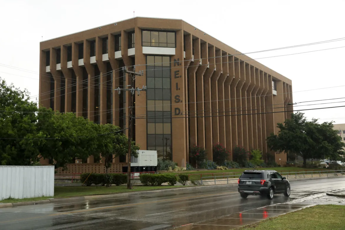 Exterior of the North East Independent School District (NEISD) Administrative Office, featuring a large, multi-story building with the NEISD acronym displayed prominently on the facade, and a car driving by on a rainy day.