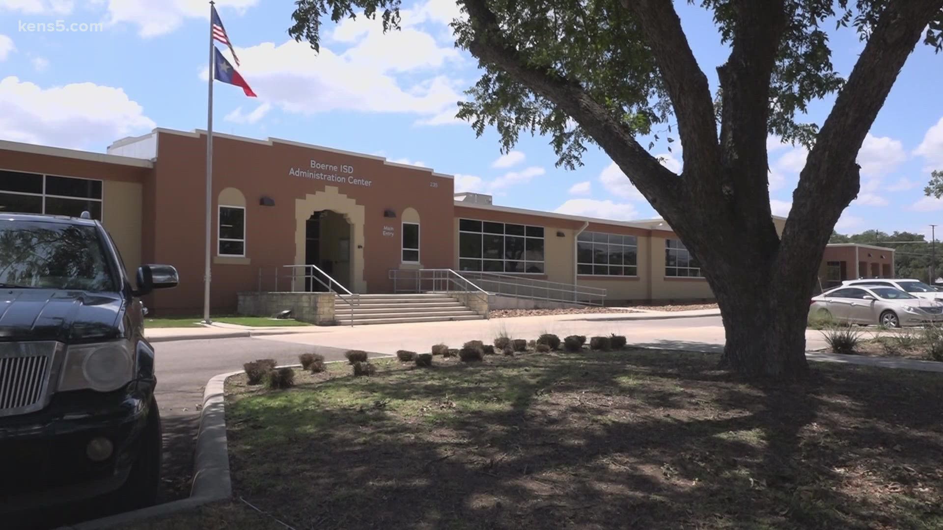 Exterior view of the Boerne ISD Administration Center, featuring the main entrance with American and Texas flags flying, a tree in the foreground, and parked cars.