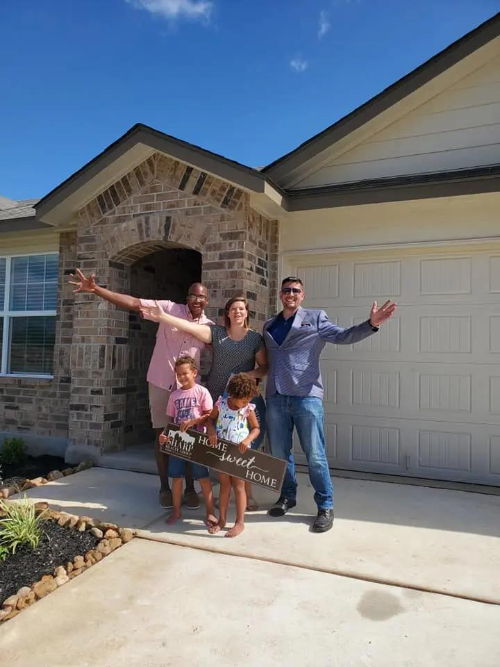 Smiling family of four stands barefoot with Anthony Sharp in front of their new brick house, all with arms outstretched; children hold a ‘Home Sweet Home’ sign on the driveway under a bright blue sky