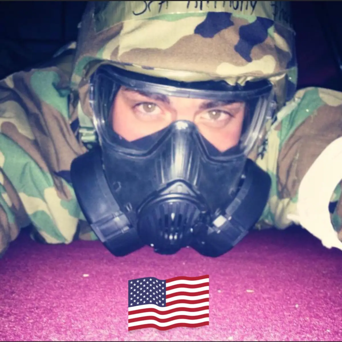 Anthony Sharp in camouflage gear lying prone, wearing a full protective gas mask and helmet, eyes looking forward; a small American flag graphic sits at the bottom of the frame