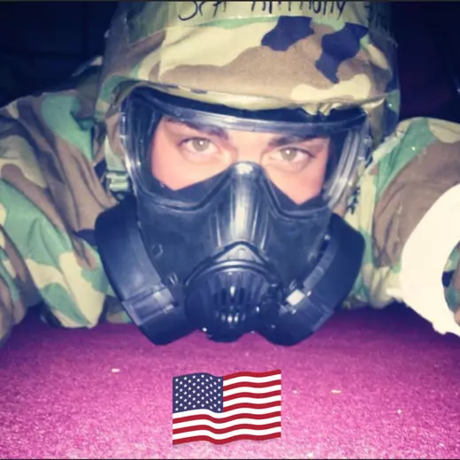 Anthony Sharp in camouflage gear lying prone, wearing a full protective gas mask and helmet, eyes looking forward; a small American flag graphic sits at the bottom of the frame