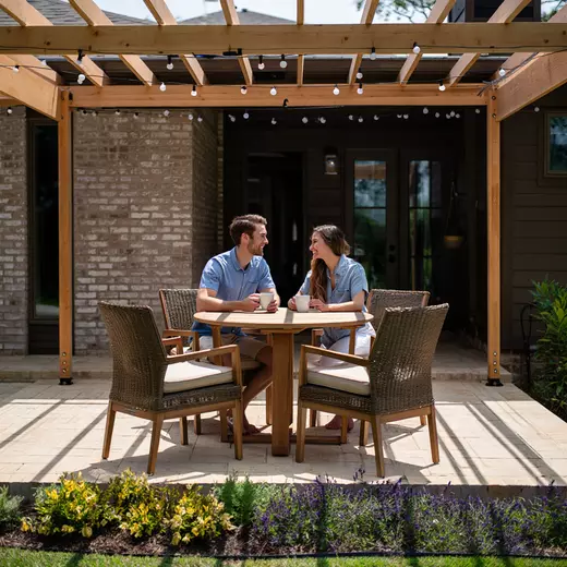 A relaxed couple in their 30s sit at an outdoor table under a pergola, sipping coffee, smiling, and enjoying a quiet morning