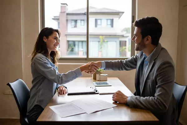 A confident buyer shaking hands with a Realtor across a negotiation table with paperwork in view