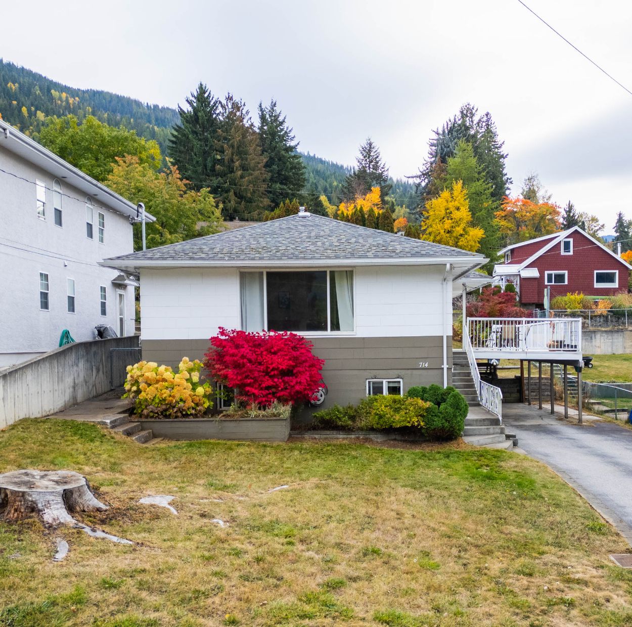 Front exterior of a 4-bedroom home in Nelson BC with mature landscaping, autumn foliage, and a long driveway leading to an elevated deck and carport