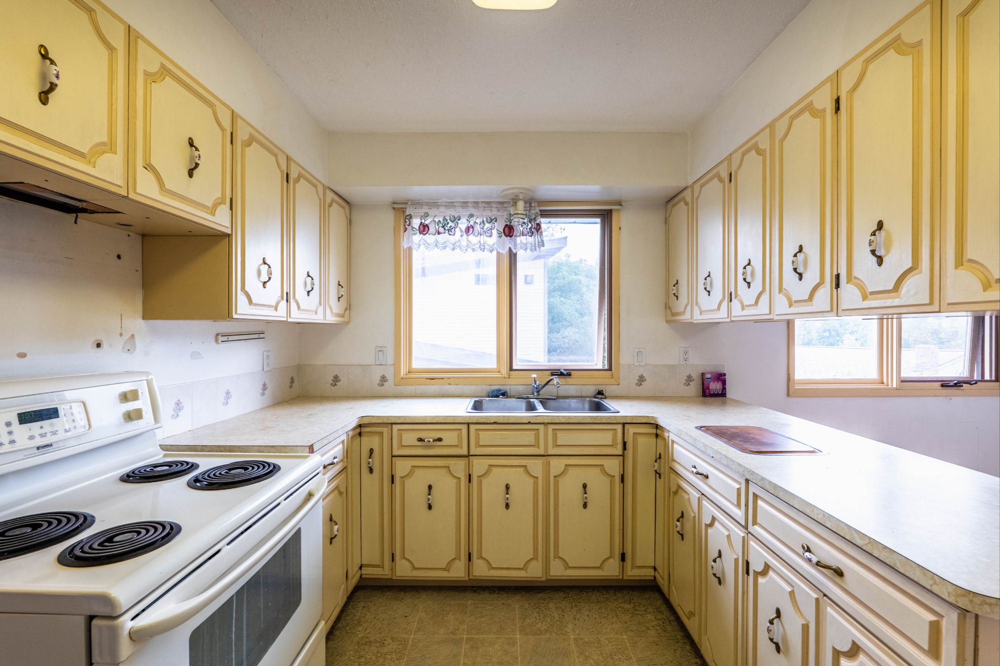 Vintage-style U-shaped kitchen with cream-colored cabinetry, electric stove, double sink, and large windows providing natural light.