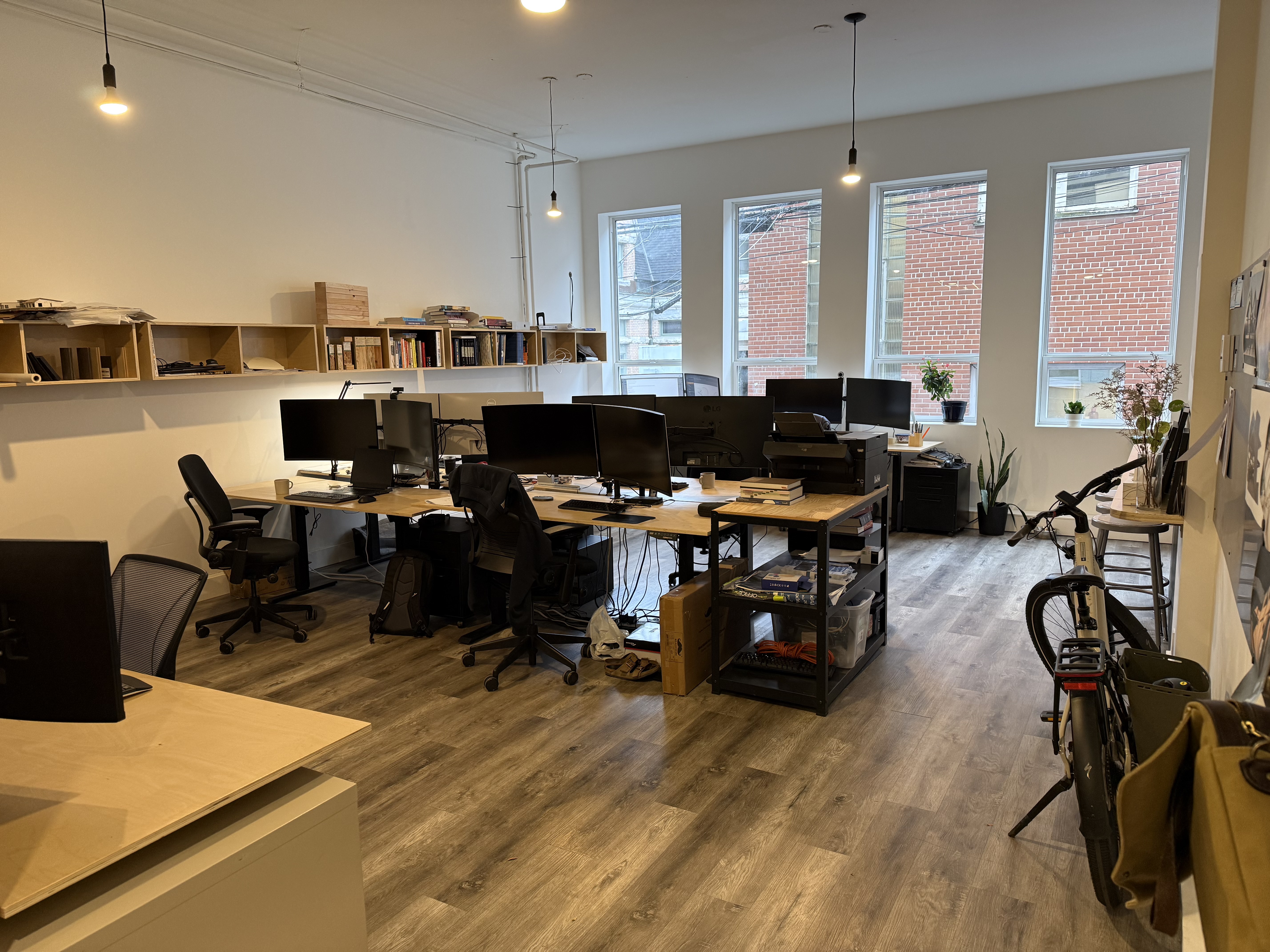 A bright, modern office with high ceilings, large windows along one wall, and soft natural light. The space features multiple desks with computer monitors, ergonomic chairs, open shelving on the back wall, and wood-look flooring. Plants and minimal décor add a professional, contemporary feel. A bicycle is parked near the front corner, emphasizing the office’s practical and flexible layout.
