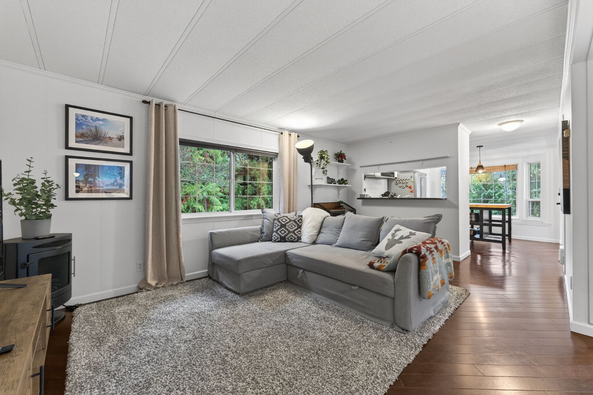 Bright and updated living room in a North Shore Nelson BC home for sale, featuring a large grey sectional sofa, modern flooring, big window overlooking lush trees, and an open-concept layout connecting to the dining area and kitchen. Cozy, move-in-ready space in a lakeside property near Kootenay Lake.