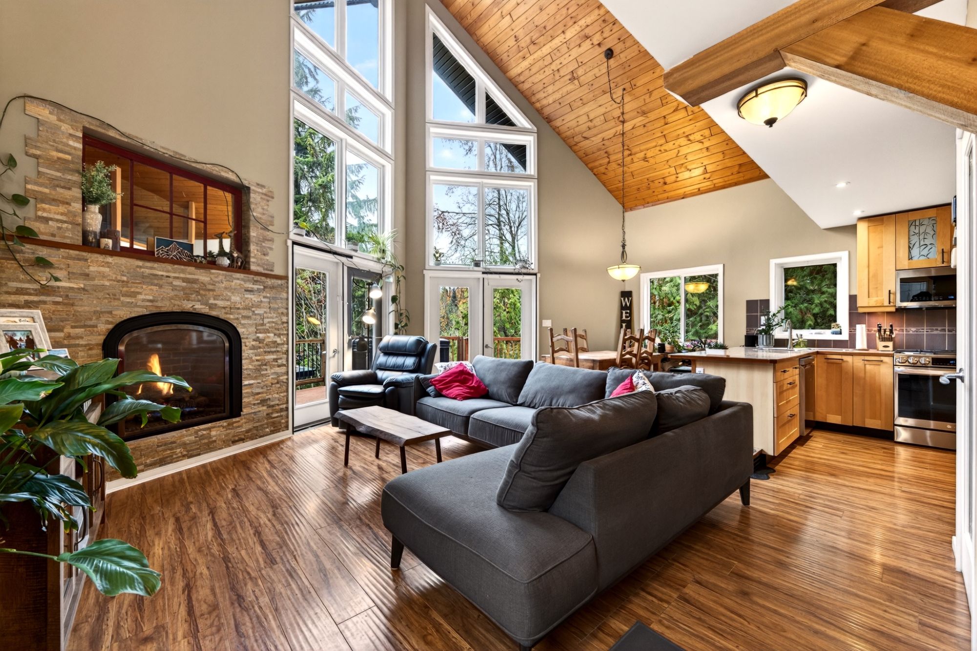 Open-concept living room with soaring vaulted ceilings, a wall of large windows, a stone fireplace, warm wood flooring, and a spacious modern kitchen and dining area in a contemporary mountain-style home.