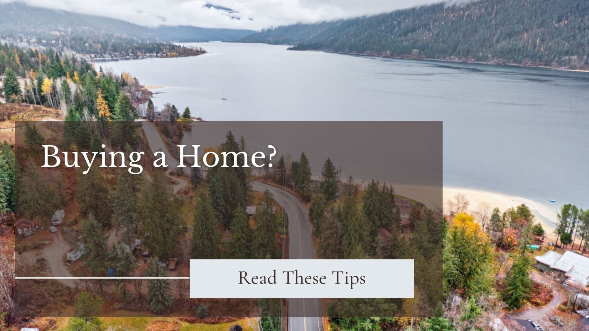 Aerial view of a winding road through a forested area beside Kootenay Lake near Nelson, BC, with mountains in the background. Graphic text overlay reads “Buying a Home?” with a button-style box saying “Read These Tips.