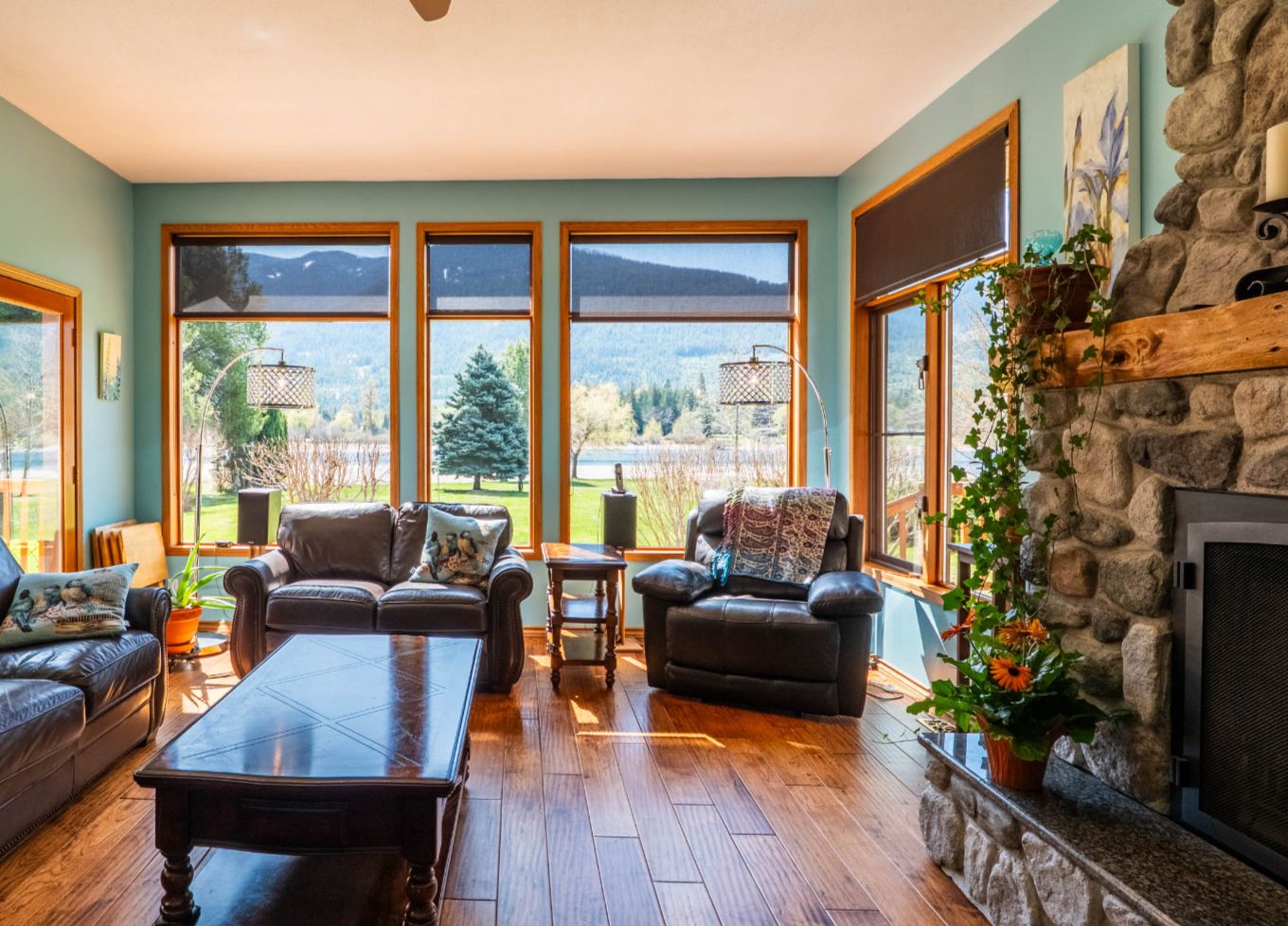 Bright living room with leather sofas, stone fireplace, and large windows offering mountain and lake views in a waterfront home near Nelson, BC.