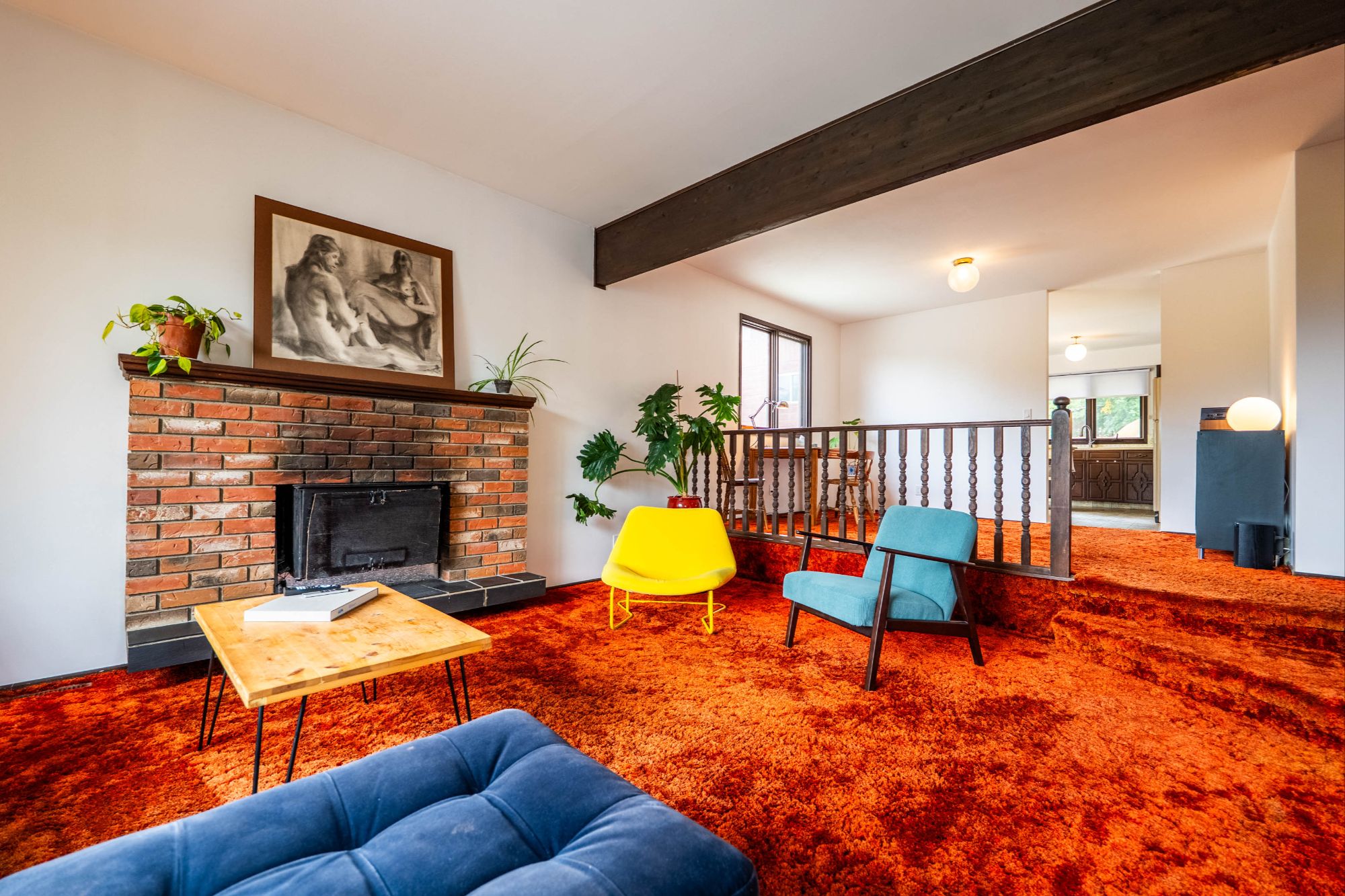 Mid-century living room featuring bold orange shag carpet, exposed wooden beam, a brick fireplace with wood-burning insert, and vintage furnishings in a split-level layout.