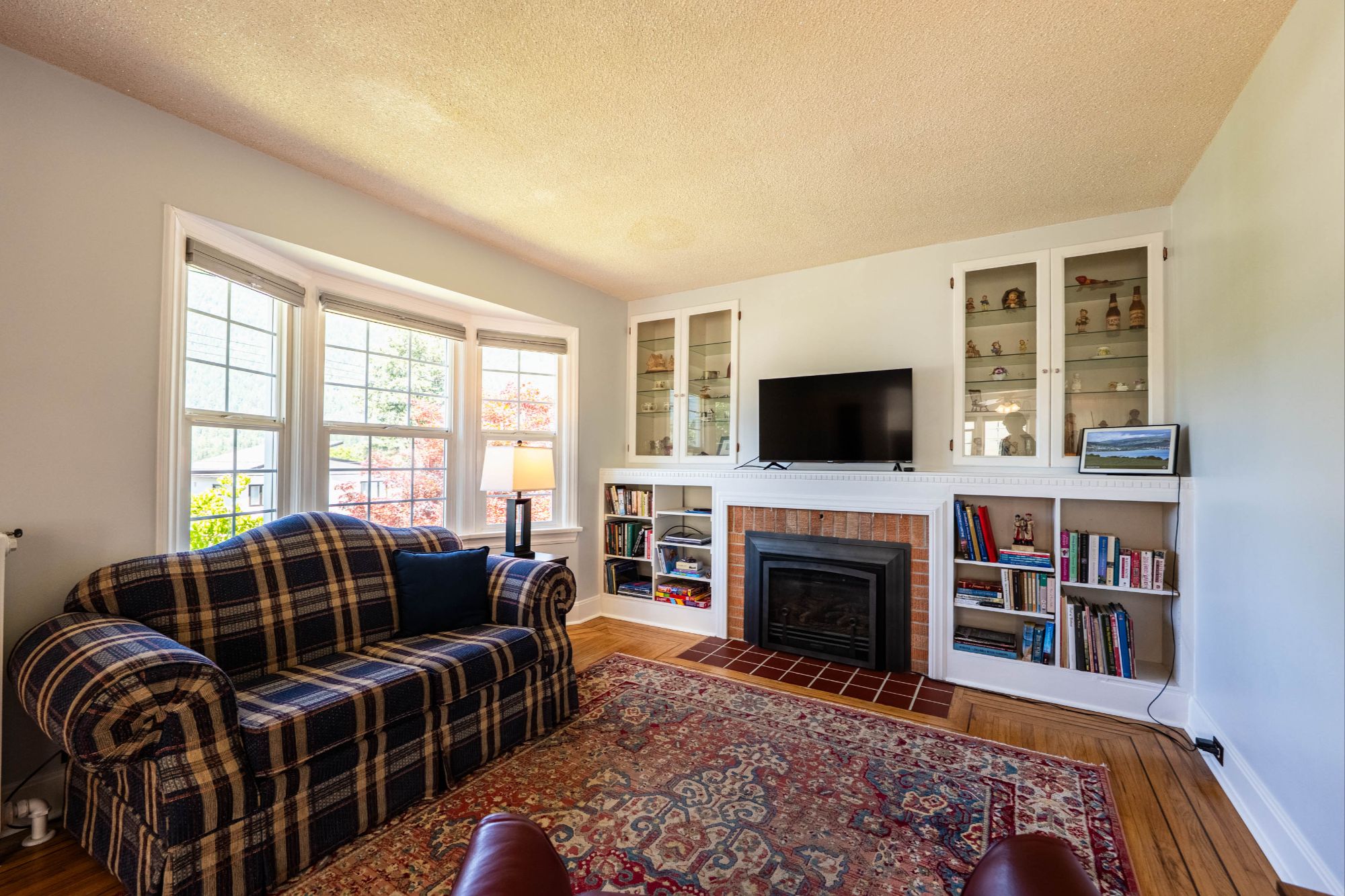 Bright and cozy living room featuring a bay window, plaid sofa, built-in shelving, gas fireplace, and warm hardwood floors at 514 Fourth Street, Nelson, BC