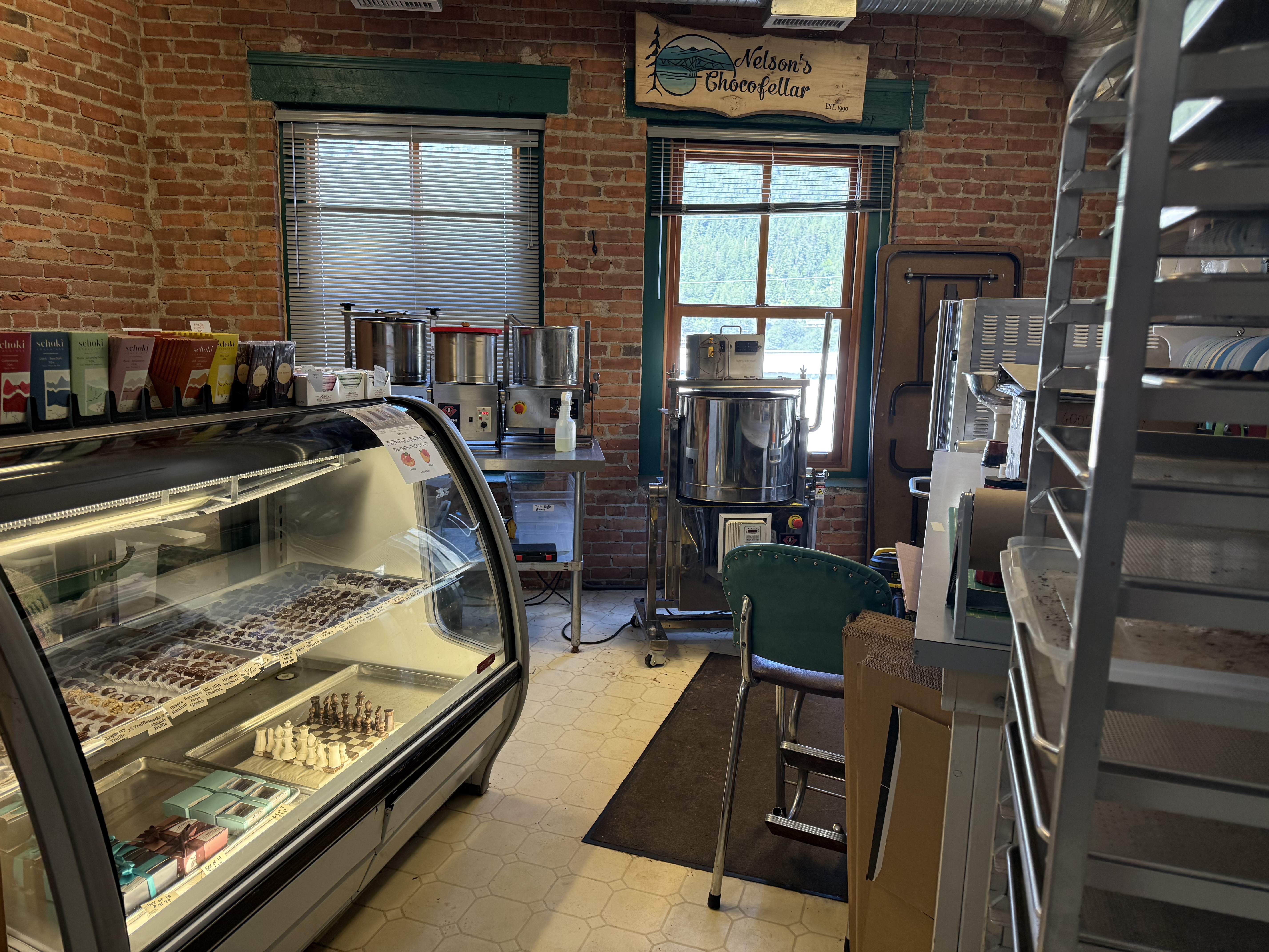 Interior of a commercial chocolate shop in Nelson, BC, featuring exposed brick walls, large windows with views of Slocan Lake, stainless steel equipment, display cases filled with chocolates, and shelving racks — located in the historic Cornerbrick Building.