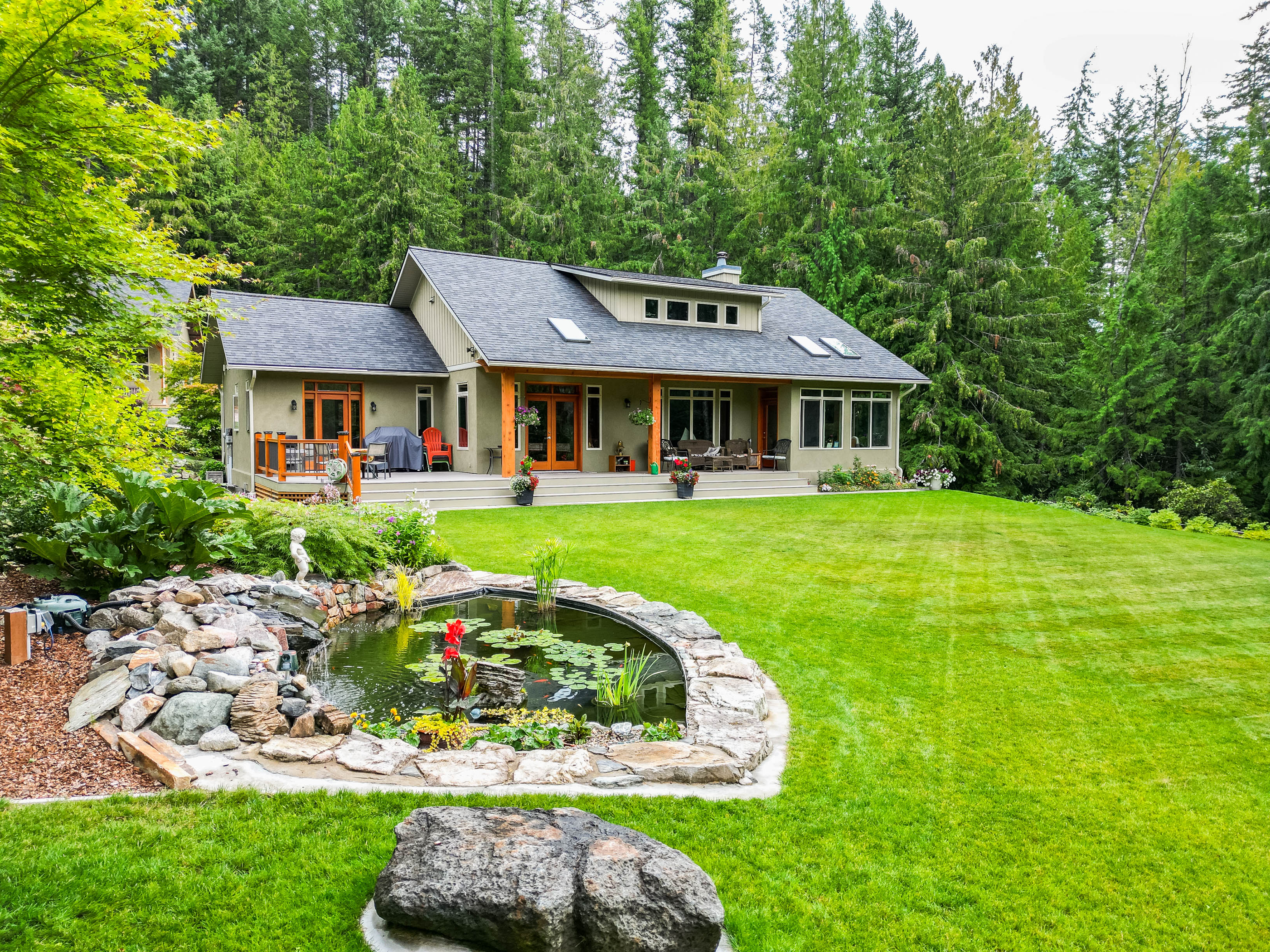 Timber frame home on 2.27 acres in North Shore Nelson BC, with landscaped lawn, koi pond, forest backdrop, and covered deck outdoor living area.