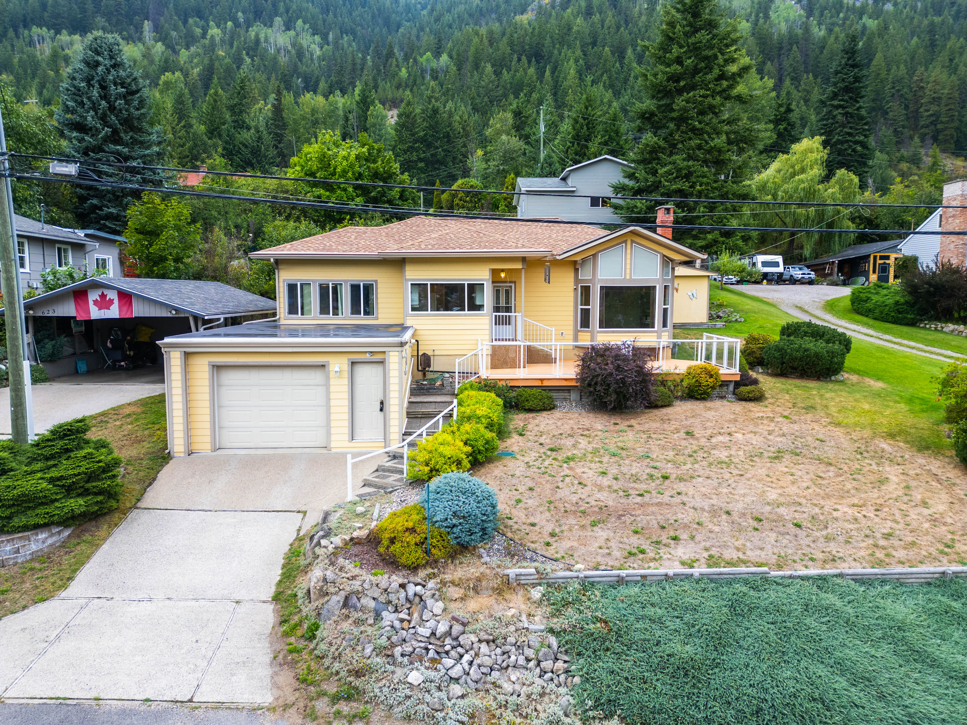 Charming yellow 2-bedroom home on Johnstone Road in Nelson BC with attached garage, landscaped front yard, and mountain backdrop.