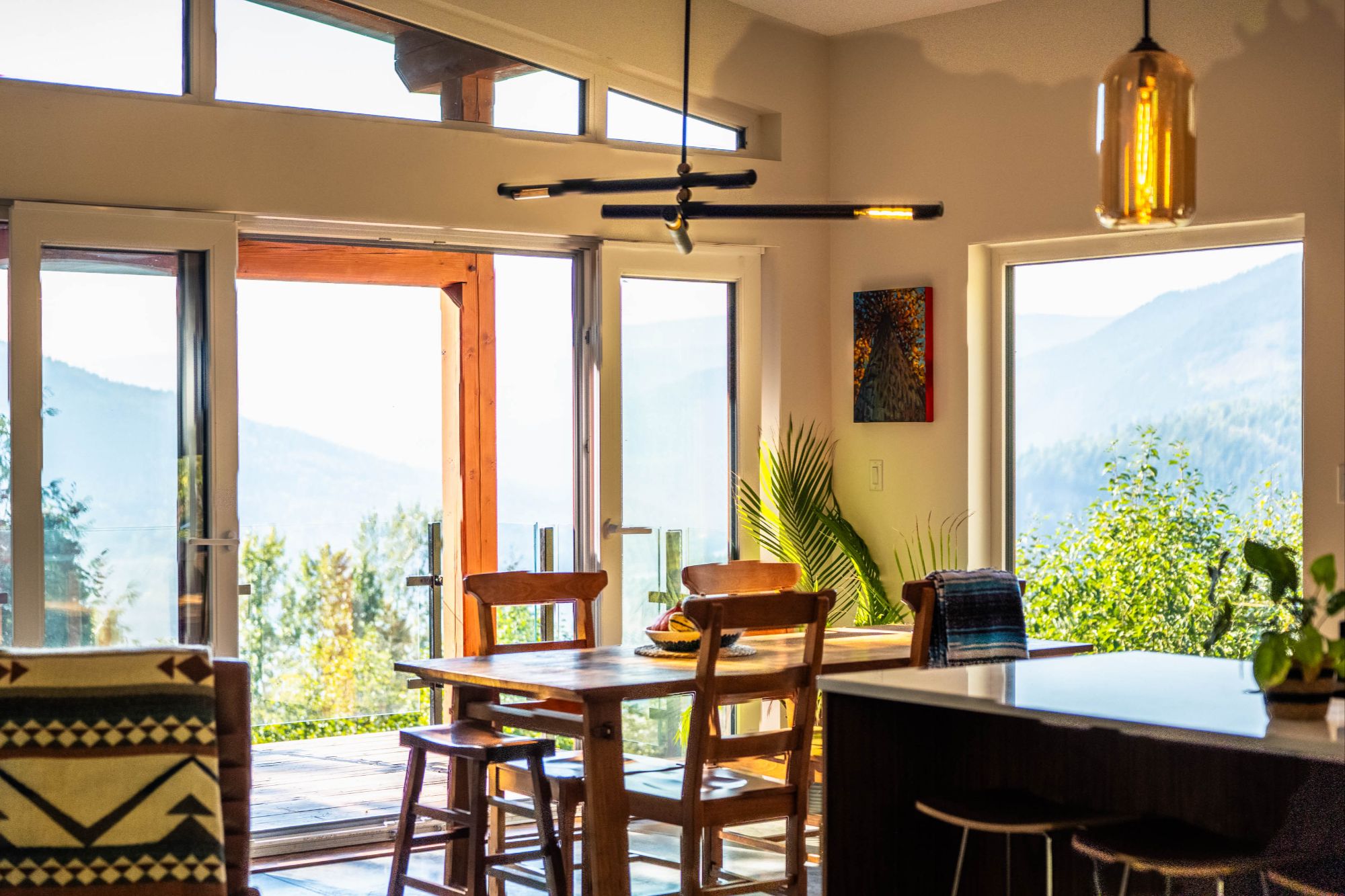 Dining area at 1409 Ash Street featuring wood furniture, modern lighting, and expansive windows with views of forested hills and mountains, opening onto a glass-railed deck in Nelson, BC"