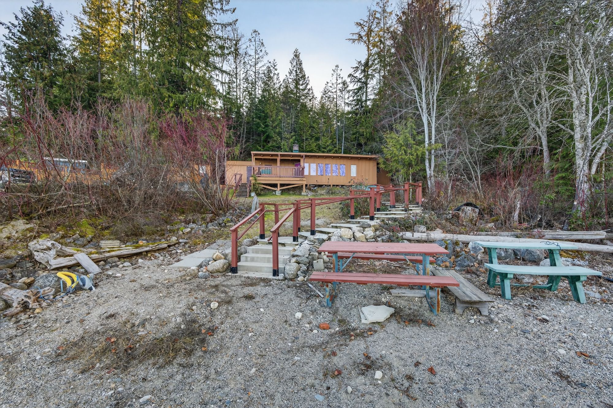 Waterfront cabin at 1294 Green Road in Queens Bay, featuring a beach-level shoreline, wooden stairs with railings leading up to the cabin, picnic tables, and forested surroundings along Kootenay Lake.