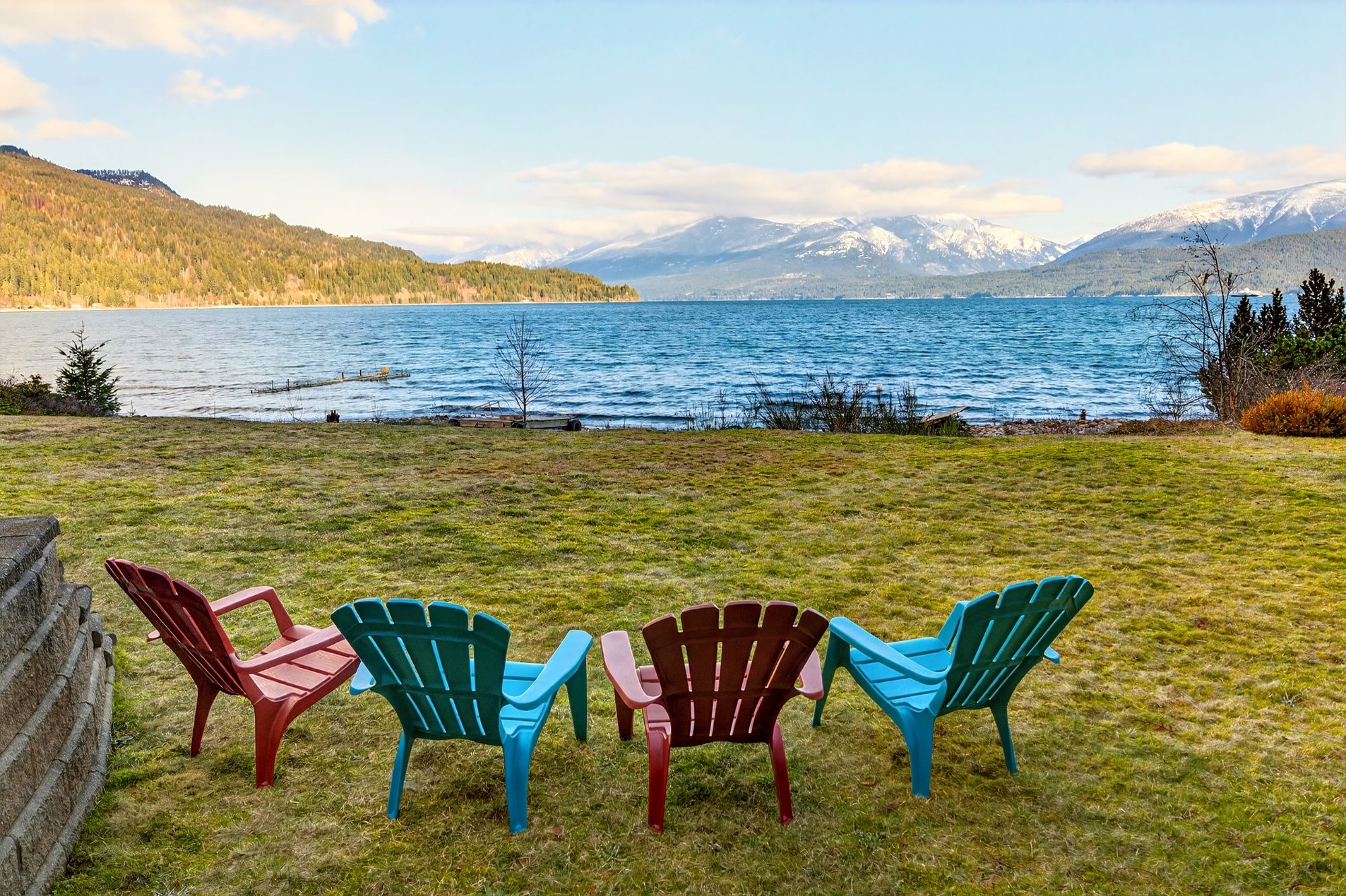 Queens Bay waterfront lawn with colourful Adirondack chairs overlooking Kootenay Lake and mountain views near Nelson, BC.