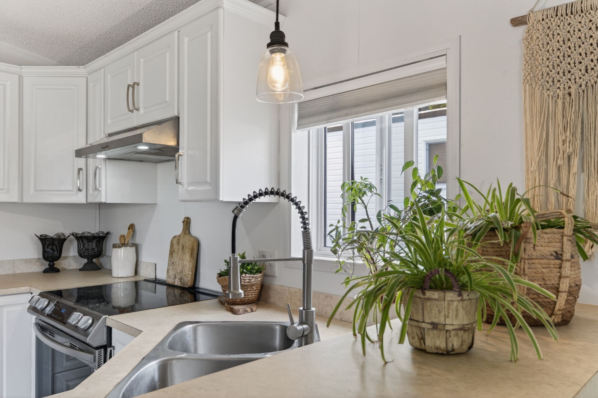 Bright kitchen at 32-1554 Granite Road in Nelson BC with white cabinetry, stainless steel stove, double sink, large window, and warm plant-filled decor.