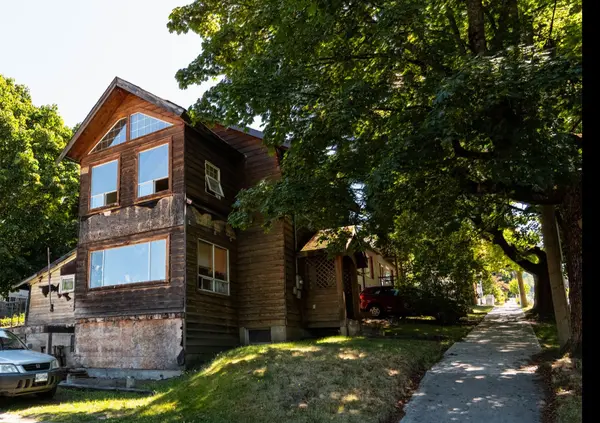 Two-storey wood-sided home in Nelson BC’s Uphill neighbourhood, shown from the street with mature trees, a sidewalk