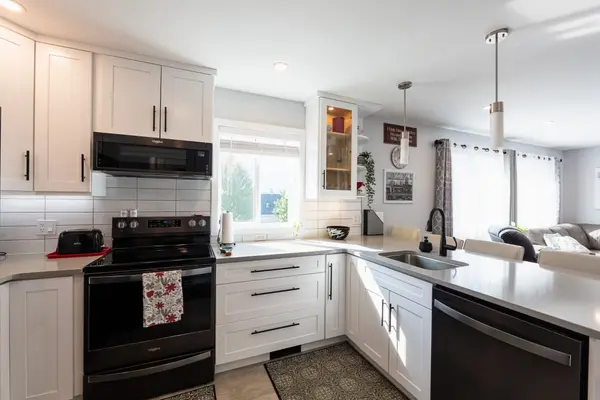 Interior view of a modern open-concept kitchen and living space in a Nelson BC home, featuring white shaker cabinets, black Whirlpool appliances, quartz countertops, pendant lighting, and light wood-style flooring flowing into a bright, spacious living area with natural light and contemporary décor