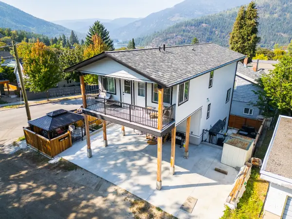 Aerial view of a modern two-storey home in Nelson BC’s Uphill neighbourhood, featuring an upper-level balcony with mountain and lake views, a large concrete driveway with overheight carport, and a private fenced patio area with gazebo — ideal for multi-family living or rental income potential.
