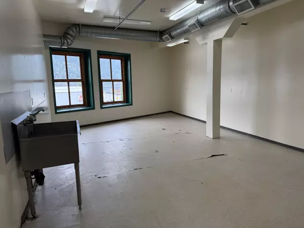 Open commercial space in Nelson’s Cornerbrick Building with two large windows offering mountain views, exposed ductwork ceiling, utility sink, and neutral walls—bright and versatile for office or retail use.