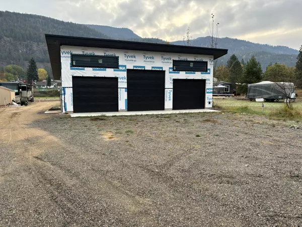 A three-bay insulated garage under construction on a semi-waterfront lot at Christina Lake, BC. The structure features black garage doors, Tyvek house wrap, and a modern design. The gravel driveway leads to an open lot with mountain views in the background, offering a peaceful setting for a future home or retreat.