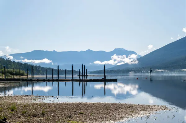 Tranquil lake view with a private dock, calm water reflecting surrounding forested mountains under a clear blue sky.