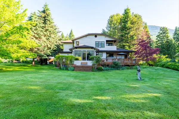 Two-storey home with wood siding and a large wraparound deck, surrounded by a manicured lawn, mature trees, and mountain views in the background.