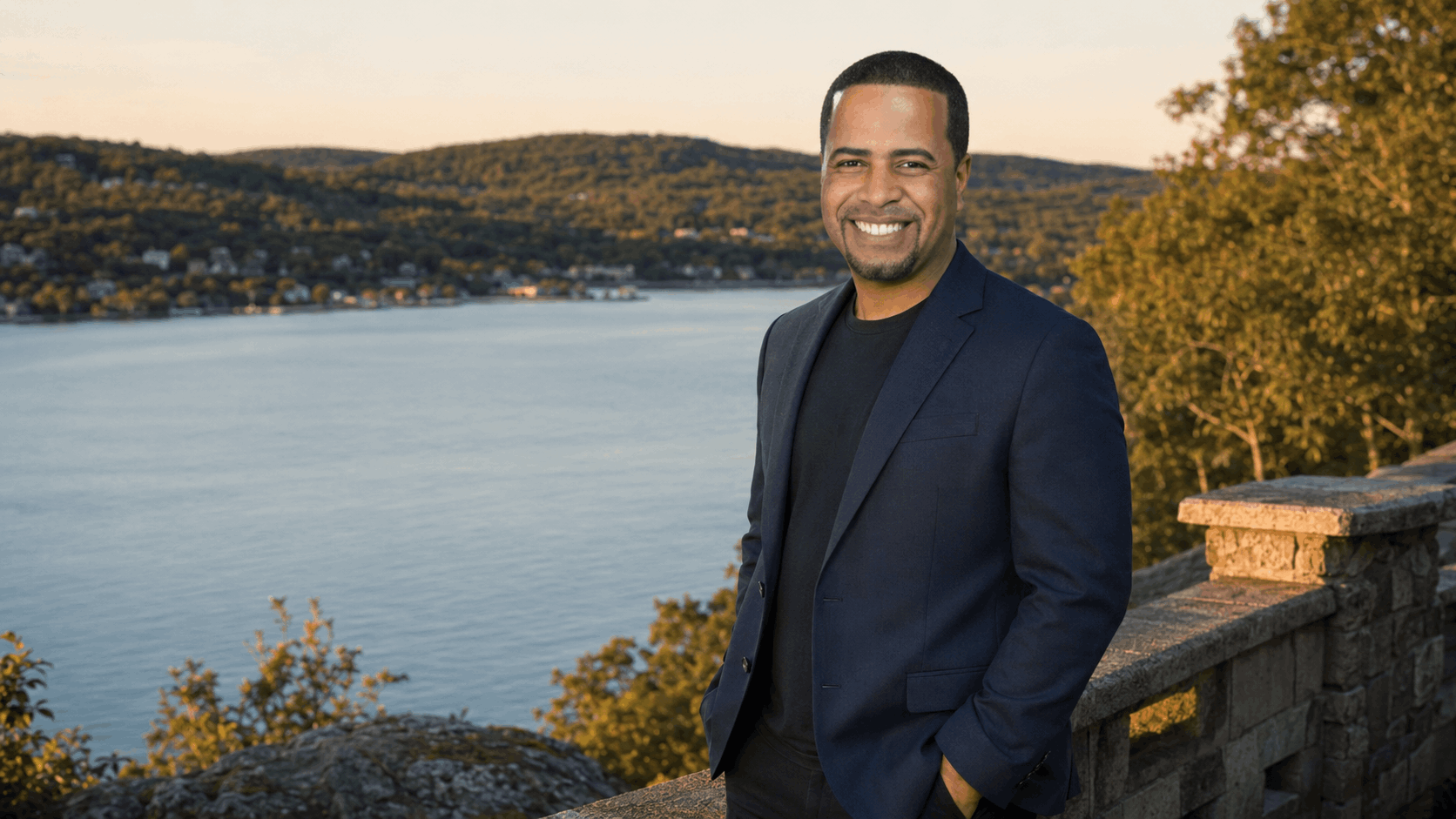 Confident Latino real estate professional standing at a North Jersey lakeside overlook with calm water, wooded hills, and golden-hour light.