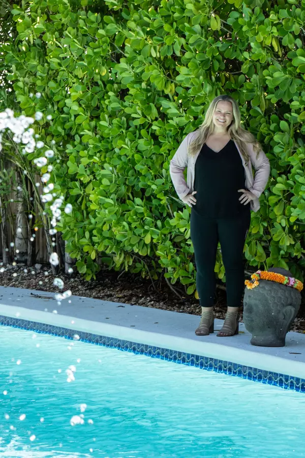 Florida Keys realtor with blonde hair dressed professionally, standing behind a teal pool with a fountain, surrounded by lush green vegetation and vibrant celosia plants in the background