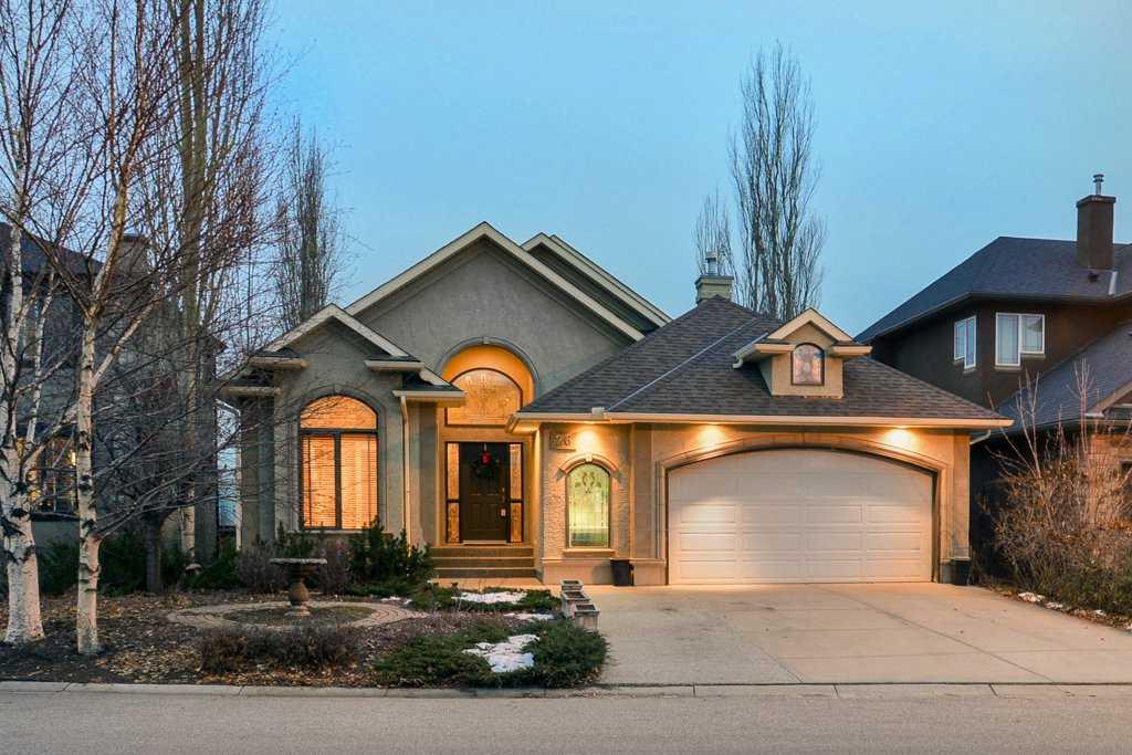 Stucco bungalow with an arched front entry and double garage at dusk, warm light in the windows, bare birch trees and patches of early snow on the front yard.