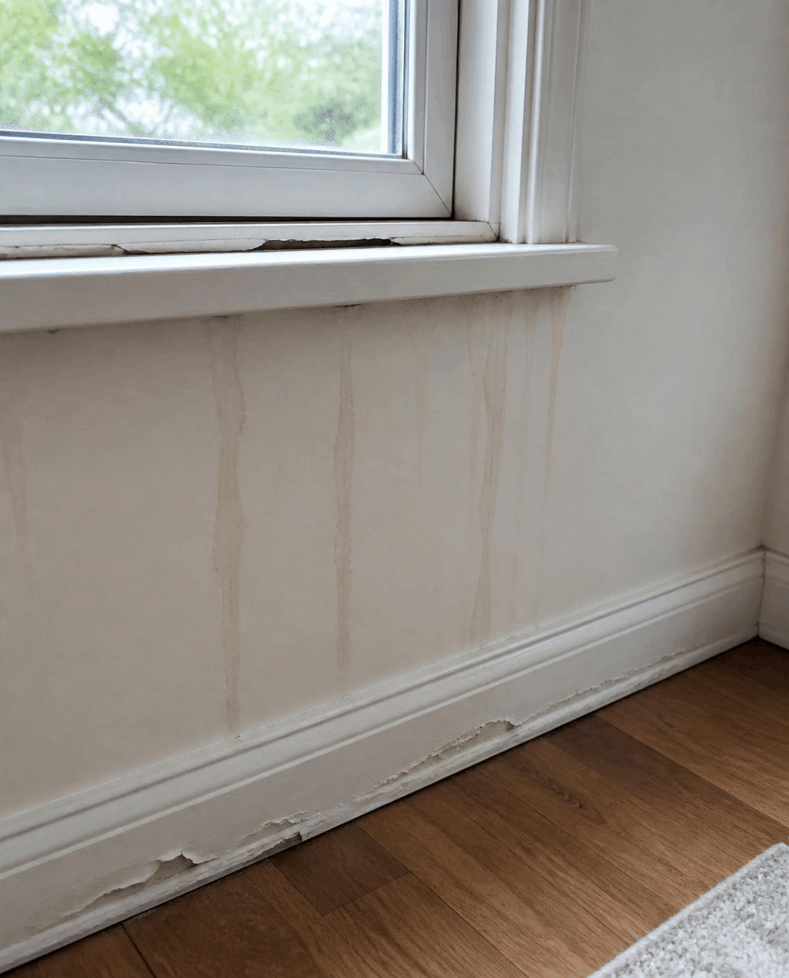 Window sill with separated caulking, faint water marks on the wall below, and a swollen baseboard with flaking paint.