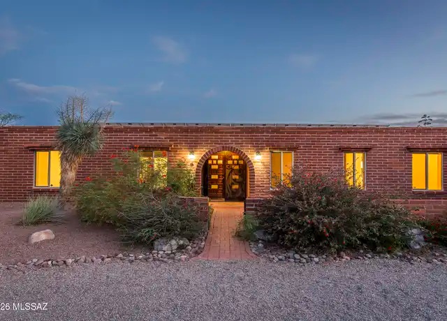 Long, low burnt adobe brick ranch home at dusk with an arched entry framing a carved wood door, warm lit windows along the facade, a brick walkway, gravel landscaping, and a yucca at one corner.