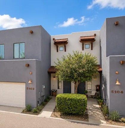 Two story attached Tucson townhomes in gray stucco with wood corbels and a tile accent roof above the entries, unit numbers 5102 and 5104, an attached garage, and a small tree in the shared front landscaping.