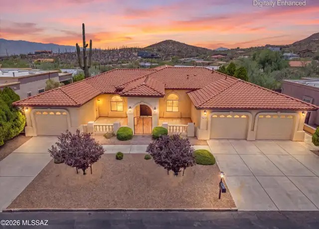 Spanish Mediterranean style Tucson home at twilight with a red clay tile roof, an arched entry gate flanked by a balustrade, arched garage details, and desert foothills and a saguaro behind it.