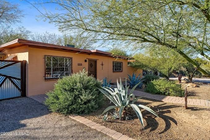 Single story Tucson bungalow with painted brick walls, a low pitched roof with wide eaves and red trim, decorative iron window grilles, and a front yard of agave, gravel, and a palo verde tree.