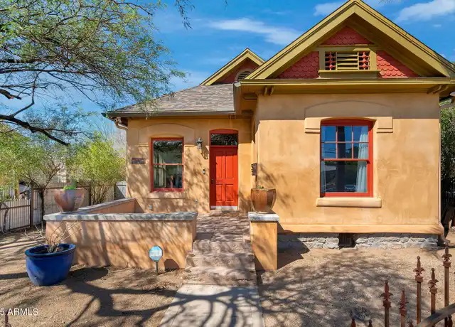 Small Victorian era Tucson cottage painted gold with red trim, a red front door, arched windows, a decorative gabled dormer with red shingles, and a low stucco garden wall shaded by a mesquite tree.