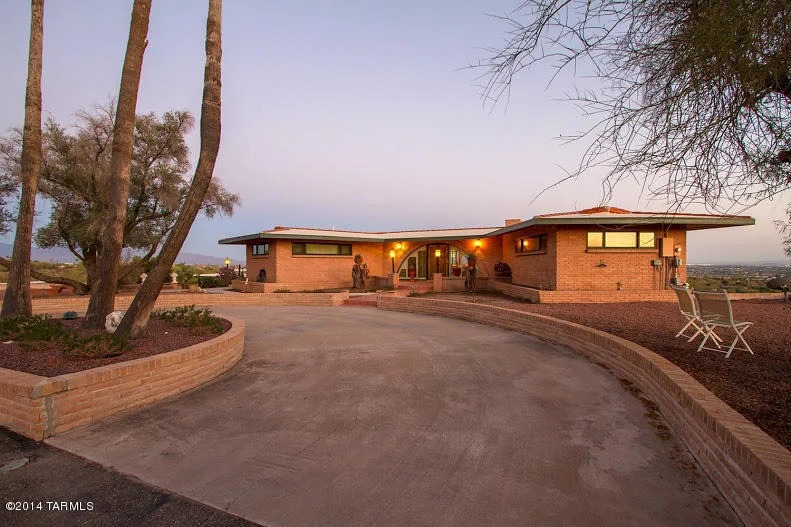 Mid century modern brick home with strong horizontal lines and deep roof overhangs, a curved brick planter wall wrapping a circular drive, mature trees, and a panoramic city view at twilight.