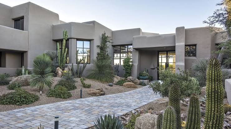 Contemporary Pueblo Revival home in Tucson with stepped flat roofs, parapet walls, and smooth stucco exterior, fronted by desert landscaping with saguaros, ocotillo, agave, and a stone paver walkway.